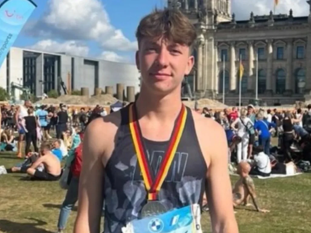 Young man wearing a medal and sportswear stands outdoors at an event with other people and historic buildings in the background.